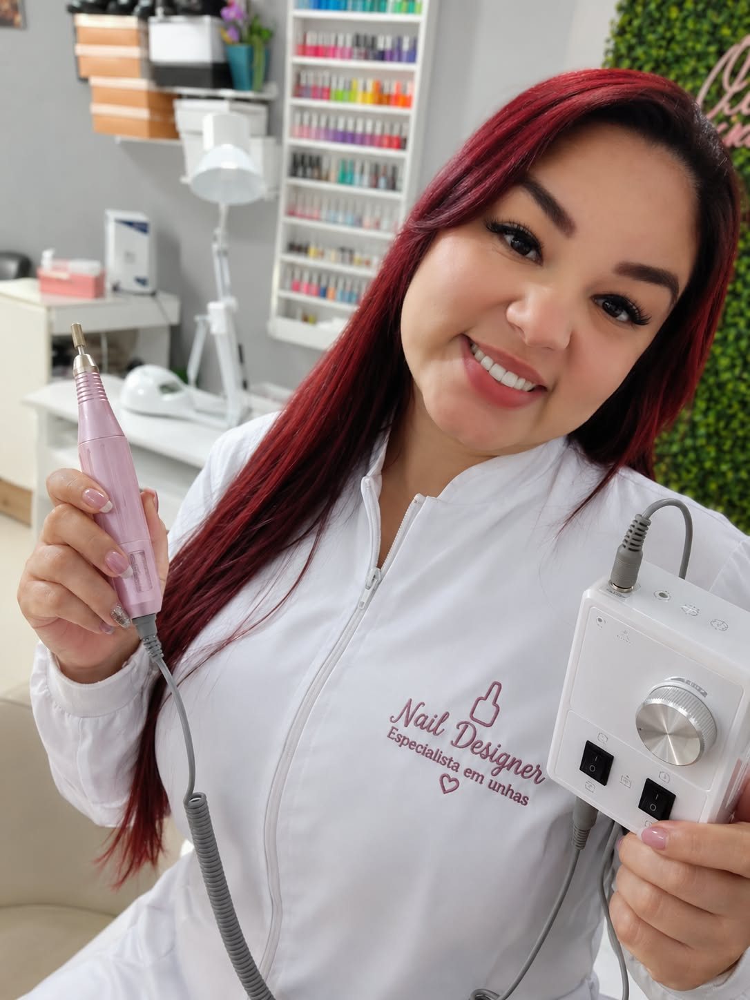 Gelise sorrindo, de jaleco branco de nail designer, segurando o motor de unhas rosa no seu espaço de atendimento